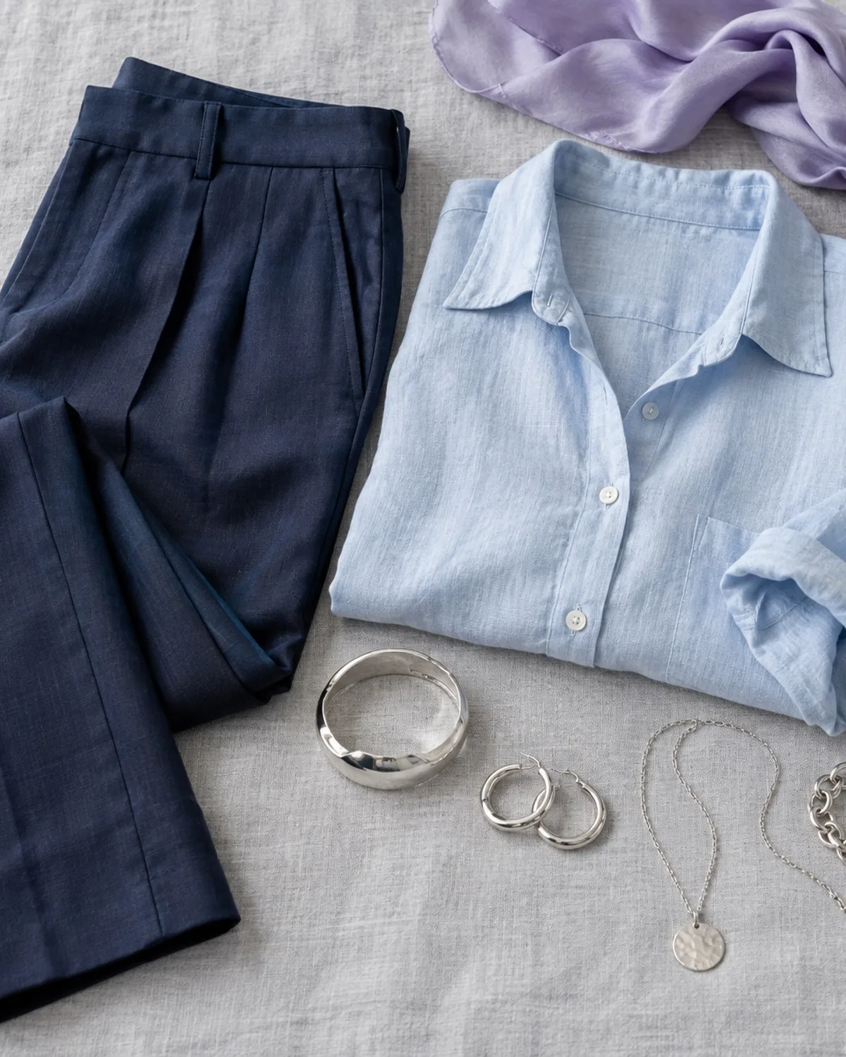 Light Summer workwear outfit flat lay: powder-blue shirt, soft navy trousers, light grey cardigan, silver studs.